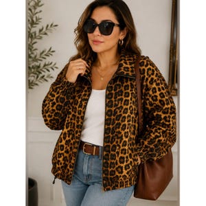 May include: A zip-up jacket featuring a brown and black leopard print pattern, worn over a plain white top with light blue denim jeans. The jacket has a high collar, long sleeves with elastic cuffs, and a brown leather belt with a gold buckle.