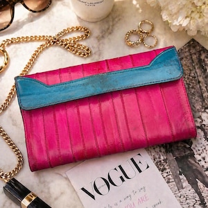 May include: A hot pink clutch with a turquoise blue flap, gold chain, and lipstick. The Vogue magazine is open to a quote about style. Sunglasses and rings are also visible.