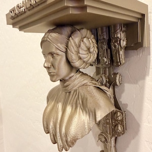 May include: A gold-colored Star Wars themed shelf featuring a bust of Princess Leia. The shelf has the Star Wars logo on the top and detailed mechanical elements on the support structure. The bust is a detailed representation of the character.