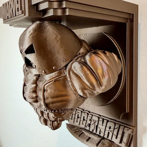 May include: A brown 3D-printed wall art piece featuring a superhero figure bursting through a circular opening. The word "MARVEL" is above the figure, and "JUGGERNAUT" is below. The figure has a metallic, textured appearance.