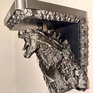 May include: A black and silver shelf featuring a detailed sculpture of Godzilla with an open mouth. The shelf has a rectangular top with the word "Godzilla" printed on the front edge. The shelf is mounted on a wall.