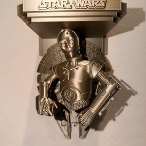 May include: A metallic gold-colored Star Wars wall shelf featuring a C-3PO figure. The shelf has the Star Wars logo at the top and is designed to hold small items. The C-3PO figure is in a classic pose.