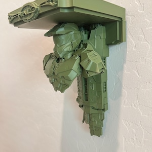 Puede incluir: Estantería verde con una figura de Master Chief de la serie de videojuegos Halo. La estantería está montada en una pared blanca y tiene el logotipo de Halo en la parte superior. La estantería mide aproximadamente 30 cm de ancho.