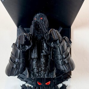 Puede incluir: Busto de Deadpool negro impreso en 3D con ojos rojos, montado en una estantería negra. El busto presenta detalles intrincados y está posicionado sobre una base con la palabra "DEADPOOL" y pistolas de juguete.