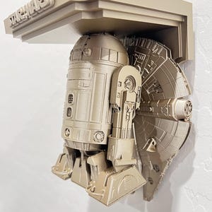 Puede incluir: Estantería de color beige con temática de Star Wars que presenta a R2-D2 y al Halcón Milenario. La estantería tiene el logotipo de Star Wars en la parte superior. La estantería está montada en una pared blanca.