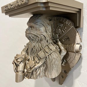 Könnte beinhalten: Beige Star Wars-Regal mit Chewbacca-Büste und Millennium Falcon-Design. Das Regal ist 3D-gedruckt und hat das Star Wars-Logo oben. Es ist für die Wandmontage konzipiert.