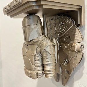 Puede incluir: Estantería plateada metálica con temática de Star Wars, con una figura de Mandaloriano y un diseño del Halcón Milenario. La estantería tiene el logotipo de Star Wars en la parte superior. La estantería está montada en una pared.
