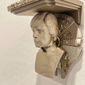 Könnte beinhalten: Beigefarbenes Star Wars Regal mit einer Büste von Luke Skywalker und einem Millennium Falcon-Design. Das Regal ist an der Wand befestigt und hat das Star Wars-Logo oben.