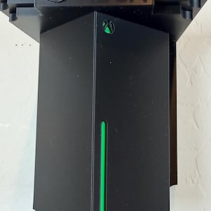 May include: Black Xbox console stand with green detailing. The stand has the Xbox logo at the top. The console is black with a green vertical stripe and green trim. The stand is designed to hold the Xbox console.