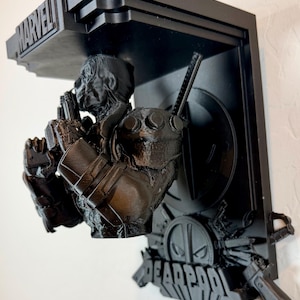 Puede incluir: Estantería de pared negra impresa en 3D con una figura de Deadpool. La estantería incluye el logotipo de Marvel y el logotipo de Deadpool. La figura sostiene dos pistolas y tiene una espada en la espalda. La estantería está montada en una pared blanca.