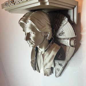 May include: A silver-colored Star Wars themed wall shelf featuring a bust of Han Solo and a Millennium Falcon design. The shelf has the words "STAR WARS" across the top.