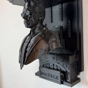 Puede incluir: Una pieza de arte mural negra impresa en 3D que presenta un busto de un hombre con gafas y un traje. La palabra "MATRIX" es visible en la pieza. La pieza de arte tiene una estructura similar a un estante sobre el busto.