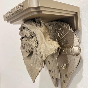 May include: A tan-colored Star Wars themed shelf featuring the Millennium Falcon and Chewbacca. The word "STAR WARS" is embossed on the top of the shelf. The shelf is mounted on a white wall.