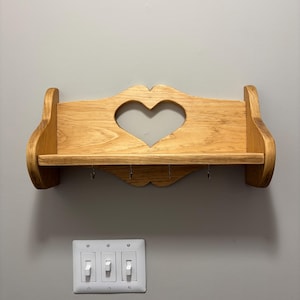 Può includere: Mensola da parete in legno con un ritaglio a forma di cuore e quattro ganci. La mensola in legno chiaro ha un design semplice e rustico. È montata su una parete grigia, con un pannello interruttori bianco sottostante.