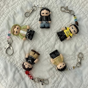 May include: Five handmade keychains with miniature figurines. Each keychain features a different figurine with unique clothing and accessories. Some keychains have beaded accents and the initials "K", "MC", "HC", and "JS". The figurines are on a white, textured surface.