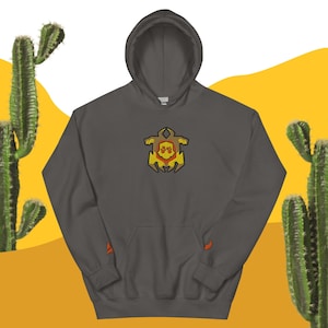May include: A dark gray hooded sweatshirt with a colorful embroidered emblem on the chest. The emblem features a geometric design with yellow, orange, and brown colors. The sleeves have small orange accents. The background includes cacti and a yellow and white backdrop.