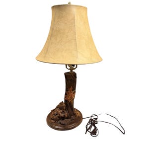 May include: A decorative table lamp with a beige lampshade and a brown, tree-trunk-shaped base. The base features carved details of animals and leaves. The lamp has a cord and plug for power.