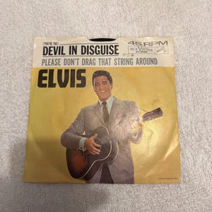 May include: Vintage 45 RPM record sleeve for Elvis Presley's "Devil in Disguise" and "Please Don't Drag That String Around." The sleeve features a photo of Elvis playing a guitar against a yellow background.