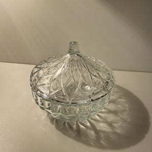 May include: A clear, round glass candy dish with a lid. The dish has a textured, geometric pattern and a small knob on top of the lid. The dish is sitting on a white surface.