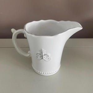 Op de afbeelding: Witte keramische kan met een handvat en tuit. De kan heeft een decoratief fleur-de-lis embleem en een kralenvoet. Het handvat is gebogen en de tuit is zacht gebogen. Waarschijnlijk voor het serveren van dranken of als decoratie.