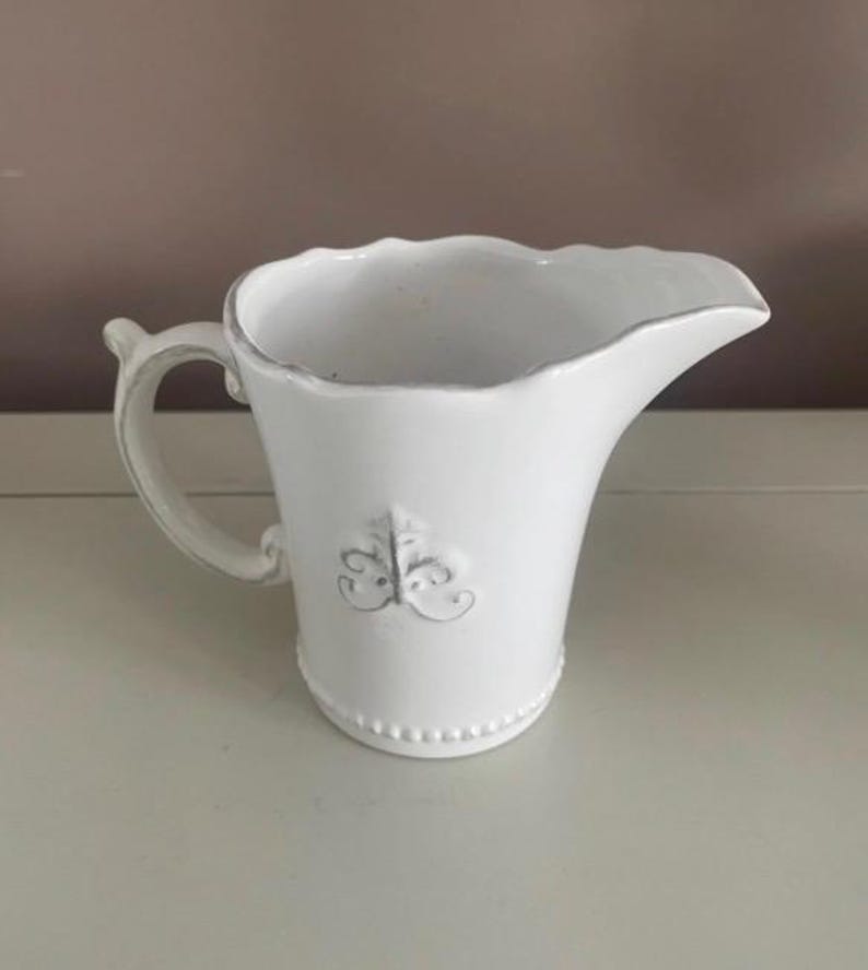 Op de afbeelding: Witte keramische kan met een handvat en tuit. De kan heeft een decoratief fleur-de-lis embleem en een kralenvoet. Het handvat is gebogen en de tuit is zacht gebogen. Waarschijnlijk voor het serveren van dranken of als decoratie.