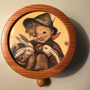 May include: Round wooden music box with a painted illustration of a smiling child in a hat, holding two white rabbits. The illustration is set against a yellow background and framed by a light brown wooden border. A small wooden knob is attached to the bottom.