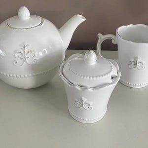 Op de afbeelding: Een witte keramische theeset met een theepot, een suikerpot met deksel en een melkkan. Elk stuk heeft een decoratief bloemmotief en een parelrand. De set is ontworpen voor het serveren van thee.