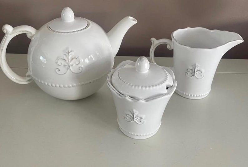Op de afbeelding: Een witte keramische theeset met een theepot, een suikerpot met deksel en een melkkan. Elk stuk heeft een decoratief bloemmotief en een parelrand. De set is ontworpen voor het serveren van thee.