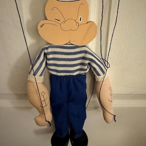May include: A Popeye marionette puppet featuring a painted wooden head with a white sailor hat, a blue and white striped shirt, blue trousers, and black shoes. The arms are made of fabric with black ink anchor tattoos and thin black control strings.