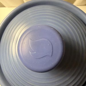 May include: A light blue circular lid with a concentric ribbed texture and a central knob featuring a raised flame-shaped emblem. The lid has a matte finish and a slightly darker blue knob that contrasts with the lighter blue surface of the lid.