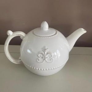 Op de afbeelding: Witte keramische theepot met een afgeronde body, tuit en handvat. De theepot heeft een decoratief reli&euml;f ontwerp aan de voorkant en een kleine knop op het deksel. Het handvat heeft een gebogen ontwerp. Een klassiek keukenartikel.