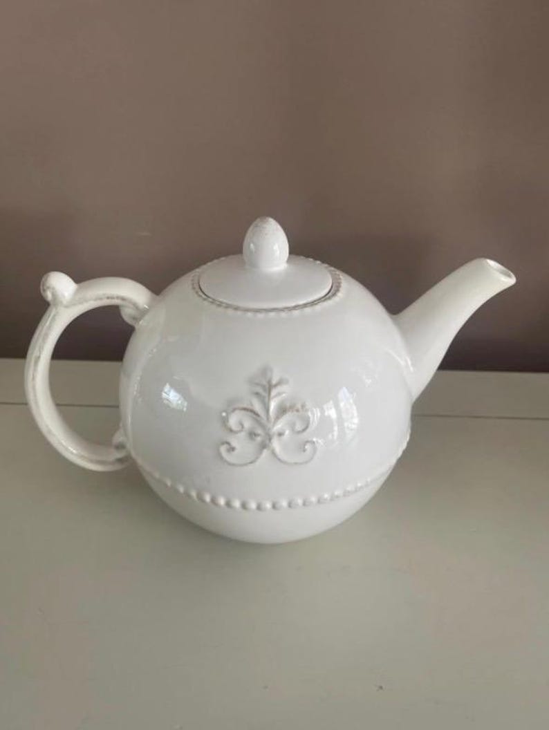 Op de afbeelding: Witte keramische theepot met een afgeronde body, tuit en handvat. De theepot heeft een decoratief reli&euml;f ontwerp aan de voorkant en een kleine knop op het deksel. Het handvat heeft een gebogen ontwerp. Een klassiek keukenartikel.