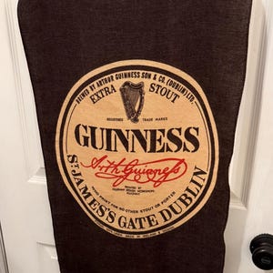 以下が含まれることがあります： ギネスのロゴが入った濃い茶色のリネンタオル。ロゴは「Guinness」の文字と「St. James's Gate Dublin」の文字が入った円形デザインです。タオルは木製のバーに掛けられており、リネン100％です。