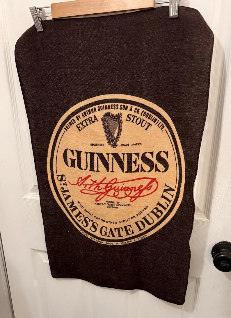 以下が含まれることがあります： ギネスのロゴが入った濃い茶色のリネンタオル。ロゴは「Guinness」の文字と「St. James's Gate Dublin」の文字が入った円形デザインです。タオルは木製のバーに掛けられており、リネン100％です。