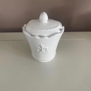 Op de afbeelding: Een witte keramische suikerpot met een deksel. De kom heeft een geschulpte rand en een decoratief reli&euml;f ontwerp. Het deksel heeft een kleine knop bovenop. De suikerpot staat op een wit oppervlak.