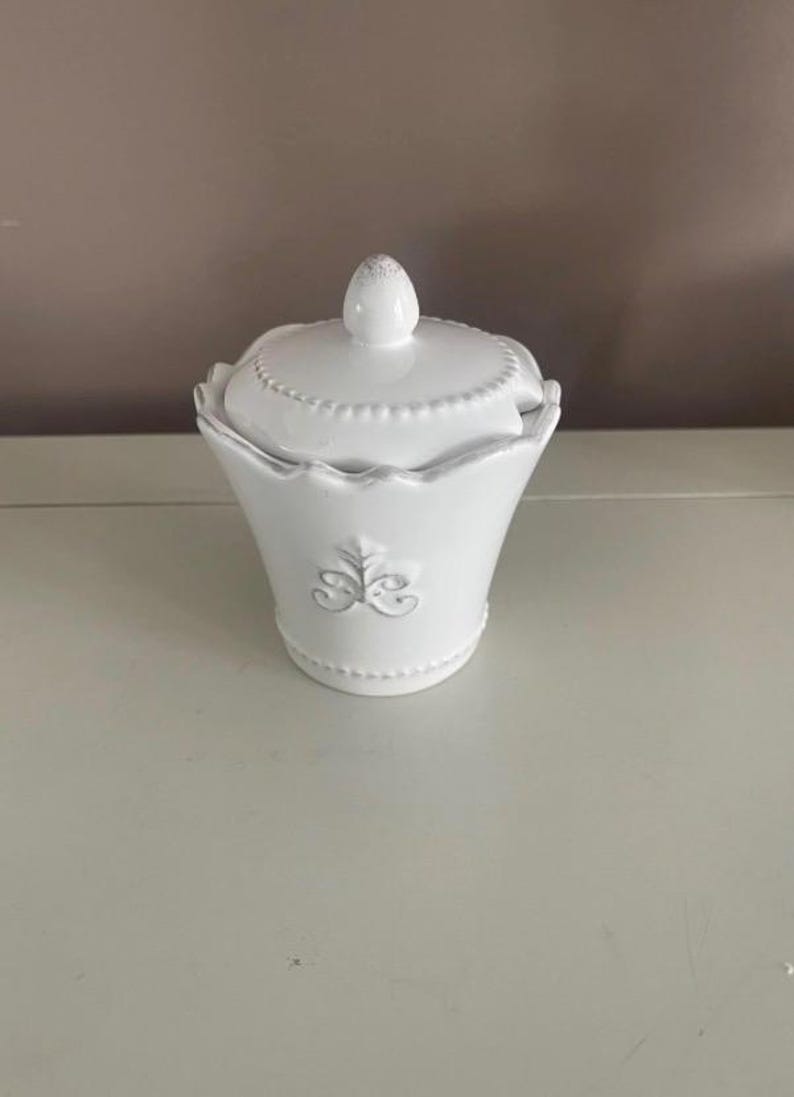 Op de afbeelding: Een witte keramische suikerpot met een deksel. De kom heeft een geschulpte rand en een decoratief reli&euml;f ontwerp. Het deksel heeft een kleine knop bovenop. De suikerpot staat op een wit oppervlak.