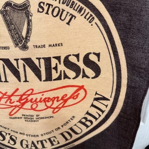 以下が含まれることがあります： ギネスのロゴが入った布製品のクローズアップ。デザインには「Guinness Stout Dublin」の文字とアーサー・ギネスのサインが含まれています。背景は暗く、ロゴは黒、ベージュ、赤です。