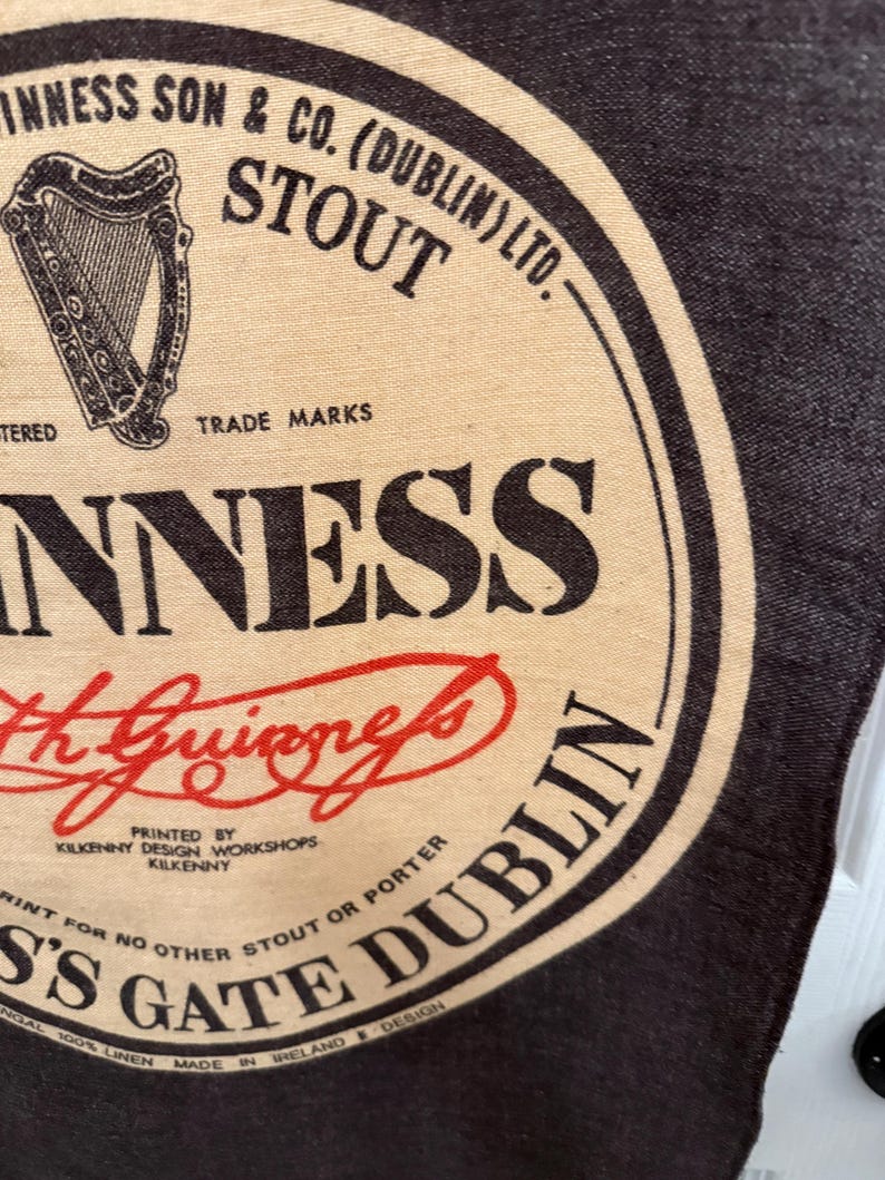 以下が含まれることがあります： ギネスのロゴが入った布製品のクローズアップ。デザインには「Guinness Stout Dublin」の文字とアーサー・ギネスのサインが含まれています。背景は暗く、ロゴは黒、ベージュ、赤です。