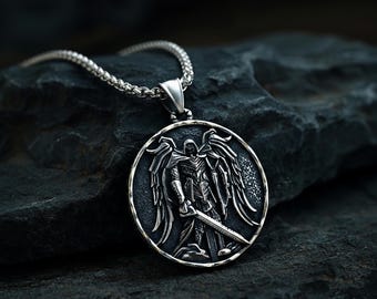 May include: A circular silver pendant featuring a detailed relief of a winged figure in armour holding a sword. The pendant has an oxidised finish to highlight the intricate design and is attached to a silver-toned metal chain necklace.