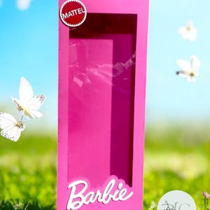 以下が含まれることがあります： 透明な窓が付いた明るいピンク色のバービーボックス。「Barbie」の文字が白い筆記体で下部に書かれています。Mattelのロゴが上部にあります。ボックスは、緑の草と青い空を背景に配置されています。