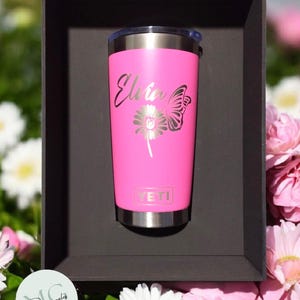 May include: A pink insulated tumbler with a silver rim and lid. The tumbler features the name "Elvia" in cursive script, along with a butterfly and flower design. The brand "YETI" is visible on the tumbler.