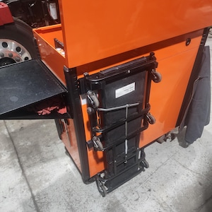 Puede incluir: Un cofre de herramientas rodante naranja con una superficie de trabajo plegable negra y un escalón plegable negro. El cofre de herramientas tiene múltiples cajones y está diseñado para uso profesional. Las ruedas facilitan la movilidad.