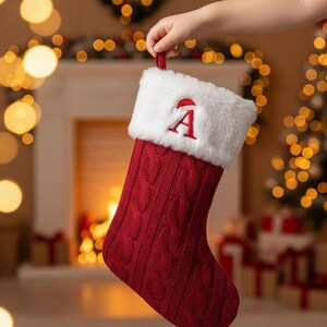 May include: A festive red Christmas stocking with a cable knit pattern. It features a white faux fur cuff and a red embroidered letter "A". The stocking is held up by a hand, with a blurred background of Christmas lights and a fireplace.