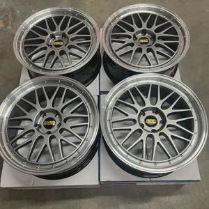 May include: Four silver alloy car wheels with a multi-spoke design. Each wheel has a silver rim and a center cap with a gold logo. The wheels are stacked on top of each other.