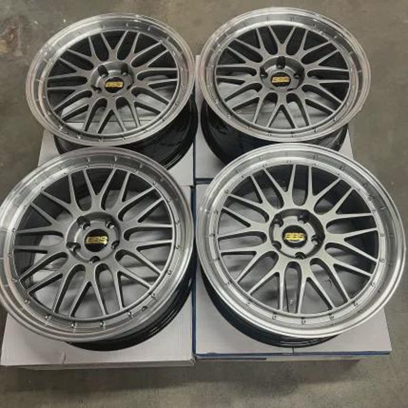 May include: Four silver alloy car wheels with a multi-spoke design. Each wheel has a silver rim and a center cap with a gold logo. The wheels are stacked on top of each other.