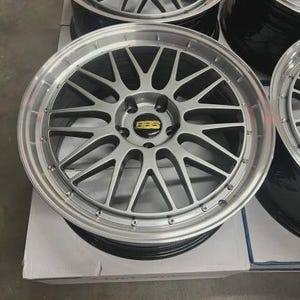 May include: Silver alloy car wheel with a multi-spoke design. The wheel has a polished silver finish with a black inner barrel. The center cap features the BBS logo in gold and black. The wheel is sitting on a white box.