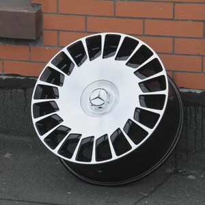 Peut inclure: Une jante de voiture noire et argent&eacute;e au design distinctif. La jante pr&eacute;sente un centre argent&eacute; avec un logo Mercedes-Benz et des accents noirs. La jante est pos&eacute;e contre un mur de briques.