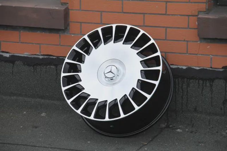 Peut inclure: Une jante de voiture noire et argent&eacute;e au design distinctif. La jante pr&eacute;sente un centre argent&eacute; avec un logo Mercedes-Benz et des accents noirs. La jante est pos&eacute;e contre un mur de briques.