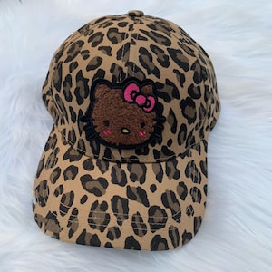 May include: A baseball cap with a tan and black leopard print pattern. The cap features a brown Hello Kitty patch with a pink bow. The cap has a curved brim and a button on top.