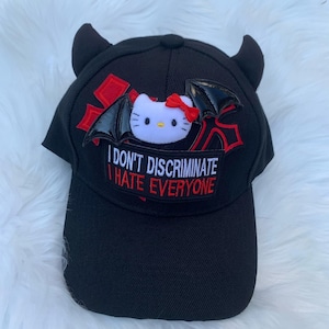 Może przedstawiać: Czarna czapka bejsbolowa z rogami diabła i naszywką z białym kreskówkowym kotem z czerwoną kokardą i skrzydłami nietoperza. Na naszywce widnieje napis "I DON'T DISCRIMINATE I HATE EVERYONE" w białym i czerwonym tekście.